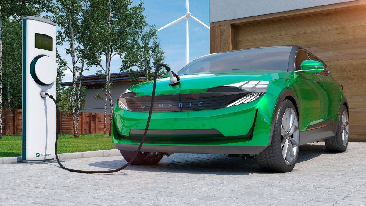 Zöld elektromos autó töltőállomáson, háttérben fák és egy ház.