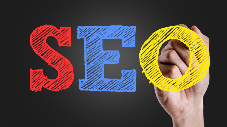 Kéz, amely színes betűkkel írja a SEO szót, háttérben sötét színnel.