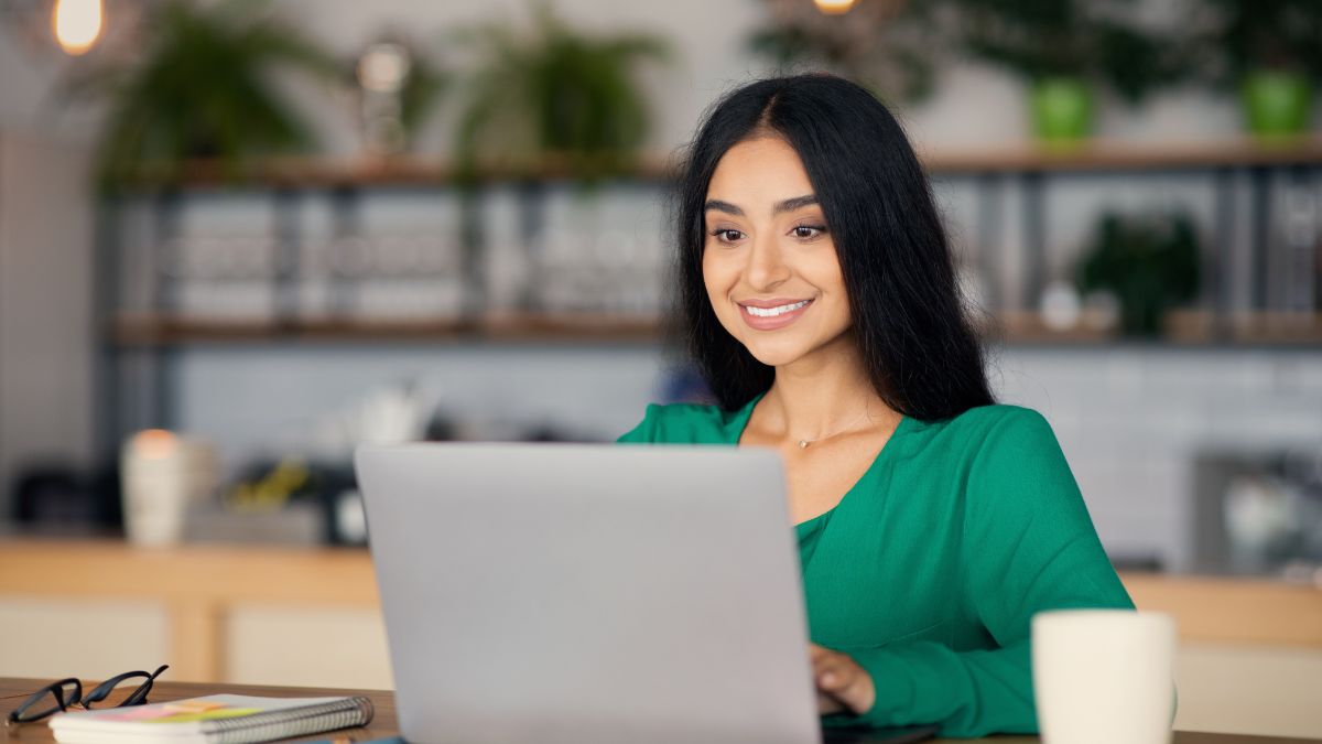 Egy fiatal nő mosolyogva dolgozik egy laptopon egy kávézóban.