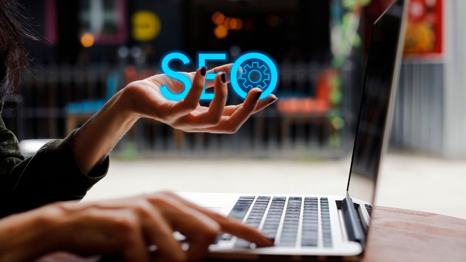 Egy női kéz, amely az SEO szót mutatja egy laptop mellett, technológiai környezetben.